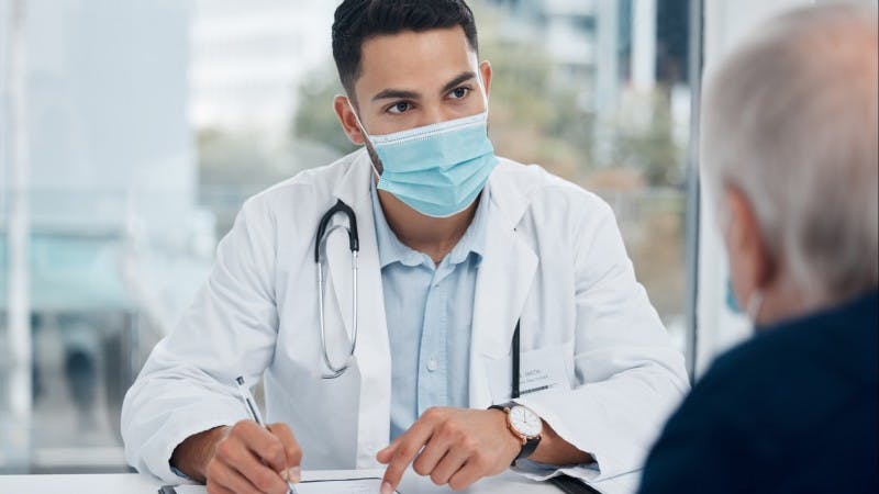 Young masked doctor talking to patient resident residency physician patient care