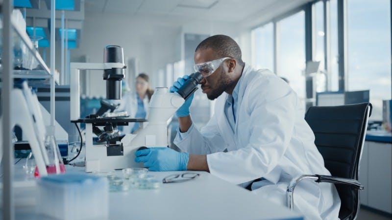 A Black, male researcher examines a specimen under a microscope in a laboratory
