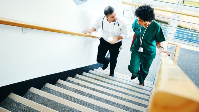 Two physicians have a conversation while walking up the stairs in a medical building.